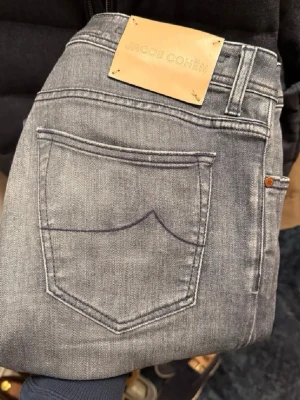 Jacob Cohën jeans - Snygga grå jeans från Jacob Cohën med klassisk femficksdesign och läderpatch bak i midjan. Jeansen har en slim passform och är tillverkade i mjukt denimtyg med lätt tvättad look. Perfekta för en trendig och avslappnad stil. 