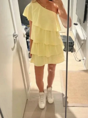 Gul one shoulder volangklänning - Snygg gul kortklänning med one shoulder-design och flera volanglager. Klänningen har en luftig och lätt känsla, perfekt för varma dagar. Passar dig som vill sticka ut med en trendig och lekfull stil.