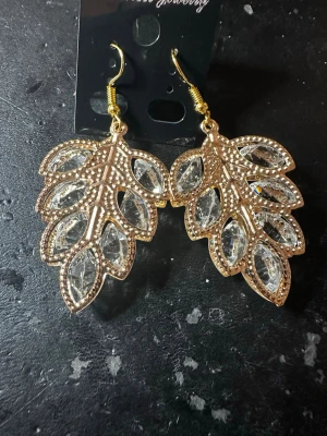 Guldiga lövörhängen med kristaller - Snygga örhängen i form av löv med guldfärgad metall och gnistrande klara kristaller. De har krokfäste och detaljerade kanter som ger ett lyxigt intryck. Perfekta för dig som vill sticka ut med accessoarer som verkligen syns.