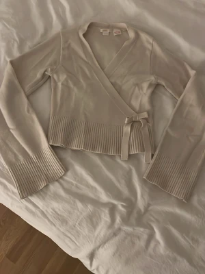 Beige omlottkofta med knytband - Snygg beige stickad omlottkofta med knytband i midjan och breda ribbade muddar vid ärmslut och nederkant. Koftan har en croppad passform och långa ärmar, perfekt att styla över ett linne eller t-shirt.