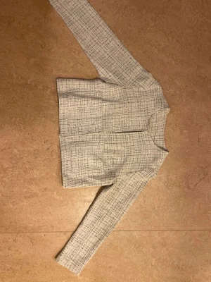 Vit tweedkofta med blått mönster - Stilren vit tweedkofta med diskret blått rutmönster. Koftan har rund halsringning, raka axlar och långa ärmar. Kortare modell utan synliga knappar eller dragkedja framtill. Perfekt för en chic och modern look. Sitter som en s eller xs.