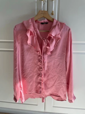 Rosa volangblus med knyt från ONLY 40 - Supersöt rosa blus från ONLY med volangdetaljer längs knäppningen och en snygg knytning vid halsen. Blusen har långa ärmar och är gjord i ett glansigt, tunt material som ger en elegant känsla. Perfekt för dig som vill ha en feminin och trendig look.