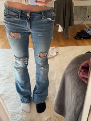 Blå slitna bootcut jeans - Säljer ett par blå bootcut jeans med slitningar och hål på låren och knäna. Jeansen har klassisk femficksmodell, låg midja och är tillverkade i denim. Perfekta för en avslappnad och trendig look.