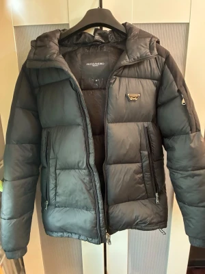 Svart pufferjacka från Alessandro Zavetti - Snygg svart pufferjacka från Alessandro Zavetti med huva, stora dragkedjeförsedda fickor och logga på bröstet. Jackan har quiltad design och dragkedja framtill samt på ärmen. Perfekt för kalla vinterdagar. Använd ett par gånger.