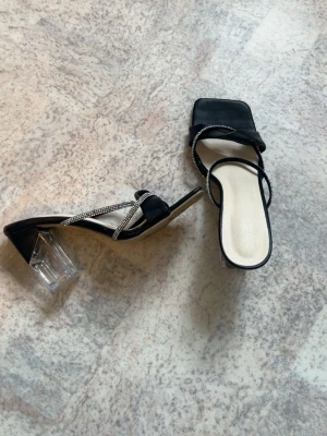 Svarta pumps med transparent klack - Stilrena svarta pumps med fyrkantig tå och transparent blockklack. Skorna har tunna remmar, varav en är dekorerad med glittrande stenar.