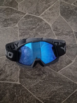 Blå spegelglas goggles YUEDI - Snygga goggles med blått spegelglas och svart ram. Justerbart svart band med YUEDI-logga i grått. Perfekta för skidåkning eller andra utomhusaktiviteter där du vill skydda ögonen mot vind och snö. Fungerar ännu bättre på moped hjälmar för att undvika motvind eller annan slags fara som kan hamna i ögonen under åkning.
