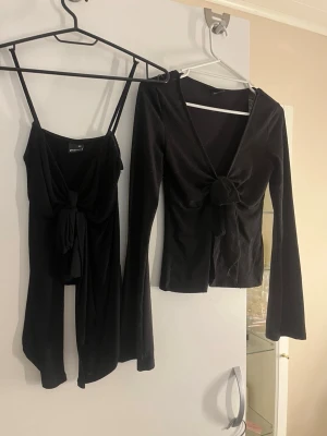 Svart omlottopp och linnetopp med knyt - Två svarta toppar från Bershka, en långärmad omlottopp med knyt framtill och en ärmlös linnetopp med knyt. Båda har djup v-ringning och smickrande passform. Perfekt för att skapa en snygg och trendig look.
