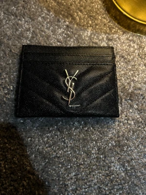 Svart kortfodral från Saint Laurent - Snyggt svart kortfodral från Saint Laurent i präglat läder med silverfärgad YSL-logga framtill. Fodralet har flera kortfack och en stilren, minimalistisk design som passar perfekt i fickan. Klassisk och tidlös accessoar för dig som gillar lyxiga detaljer.
