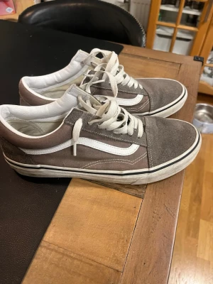 Bruna Vans Old Skool sneakers - Säljer ett par bruna Vans Old Skool sneakers med vit sula och vit sidostripes. Skorna har klassisk låg profil, snörning och ovandel i mocka och canvas. Perfekta för dig som gillar streetwear och vill ha en tidlös look.