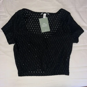 Svart croppad spetsig topp H&M - Svart croppad topp från H&M med genombruten spets och korta ärmar. Tillverkad av 52% återvunnen polyester. Toppen har en rund halsringning och sitter snyggt till höga byxor eller kjol.