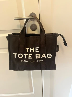 Svart The Tote Bag från Marc Jacobs - Snygg svart shoppingväska från Marc Jacobs med texten 'THE TOTE BAG' tryckt i vitt på framsidan. Väskan är tillverkad i canvasmaterial och har dubbla handtag samt dragkedja upptill. Perfekt för dig som vill ha en trendig och rymlig tote.
