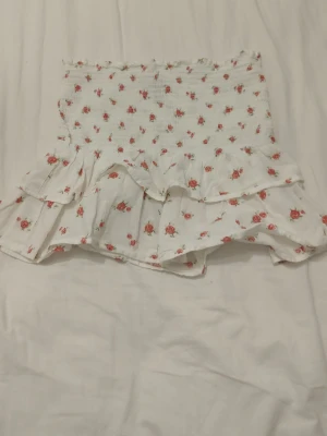 Blommig volang kjol från H&M Divided - Supersöt vit kjol från H&M Divided med små röda blommor och volangdetaljer. Smockad överdel för skön passform och dubbla volanger nedtill. Perfekt för sommarens alla tillfällen.