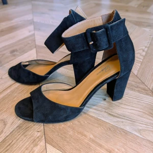 Svarta mocka-pumps med bred klack - Snygga svarta pumps från Vagabond i mjuk mocka. Skorna har öppen tå, bred klack och en bred ankelrem med stor spänne för extra statement. Perfekta för dig som vill ha både stil och komfort. Klackhöjden ger en elegant siluett och passar till många outfits.