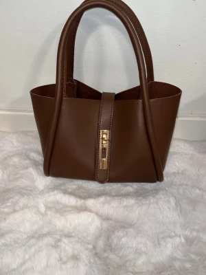 Brun handväska i läderlook - Snygg brun handväska i läderimitation med två rejäla handtag och guldfärgat spänne framtill. Väskan har en stilren, avlång form och är perfekt för dig som gillar minimalistisk design. Passar till många olika outfits och är lätt att bära med sig.