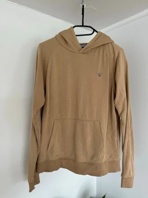 Beige hoodie från Gant med ficka - Snygg beige hoodie från Gant med klassisk känguruficka och liten broderad logga på bröstet. Mjuk bomullskänsla och avslappnad passform, perfekt för en chill look. Huva och ribbade muddar ger extra komfort.