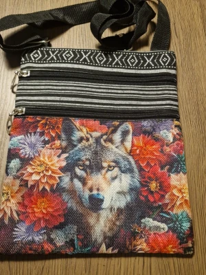 Axelväska med vargmotiv och blommor - Unik axelväska i tyg med färgstarkt vargmotiv och blommor på framsidan. Väskan har två dragkedjefack och justerbar svart axelrem. Baksidan har svartvita etnomönster. Bredd 19 cm. Höjd 23,5 cm. Perfekt för dig som gillar djur och boho-stil.