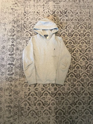 Grå hoodie från Polo Ralph Lauren - Fläcken du ser är vatten 💧 Klassisk ljusgrå hoodie från Polo Ralph Lauren med broderad marinblå logga på bröstet. Hoodien har huva, känguruficka och ribbade muddar. Tillverkad i mjuk bomullsblandning som känns skön mot huden. Perfekt för en avslappnad och stilren look.