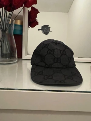 Svart keps från Gucci med GG-mönster nu model  - Snygg svart keps från Gucci med klassiskt GG-monogram över hela kepsen. Kepsen har böjd skärm och justerbar baksida. Materialet är tyg med diskret rutmönster i bakgrunden som ger en exklusiv känsla. Perfekt för dig som gillar streetwear och designeraccessoarer.