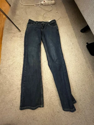 Mörkblå bootcut jeans från Lee - Snygga mörkblå bootcut jeans från Lee med klassiska gula sömmar och metallknapp med logga. Jeansen har en tidlös passform med lätt utsvängda ben och är tillverkade i slitstark denim. Perfekta för dig som gillar en retroinspirerad look.