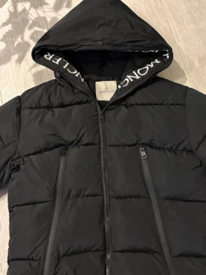 Svart dunjacka med huva från Moncler - Snygg svart dunjacka från Moncler med huva och dragkedja framtill. Jackan har två snedställda fickor med dragkedja och Moncler-logga på insidan av huvan. Perfekt för kalla vinterdagar och riktigt stilren design.