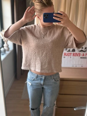 Beige stickad cropped tröja Peppercorn - Säljer en beige stickad cropped tröja från Peppercorn. Tröjan har korta ärmar och en boxig passform med ribbad kant vid halsen. Materialet är mjukt och fluffigt, perfekt för att skapa en avslappnad look.