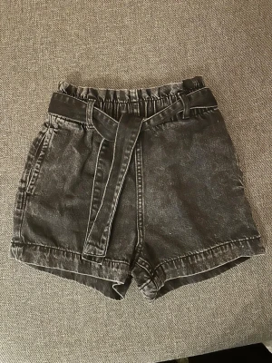 Svarta denimshorts med bälte - Snygga svarta denimshorts med tvättad look och tillhörande bälte i samma material. Shortsen har elastisk midja och två sidofickor.