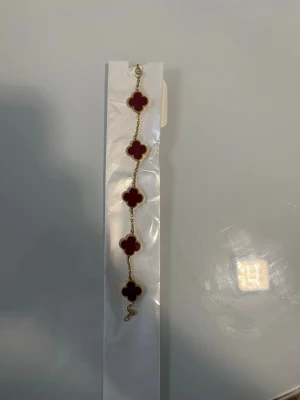 Guldarmband med röda klöver - Snyggt armband med fem fyrklöverformade hängen i röd sten och guldfärgad kedja. Klöverhänget har pärlstruktur runt kanterna som ger en exklusiv känsla. Perfekt accessoar för att lyfta din outfit med en färgklick och elegant design.