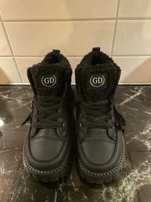 Svarta kängor med teddyfoder GD - Säljer ett par svarta kängor från GD med chunky sula och teddyfoder. Kängorna har grova snören, högt skaft och detaljer i både läderimitation och textil. Perfekta för kalla dagar när du vill ha både stil och komfort.