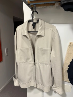 Beige overshirt från H&M - Stilren beige overshirt från H&M med två stora bröstfickor och dragkedja framtill. Overshirten har klassisk krage och är tillverkad i ett mjukt, strukturerat tyg som ger en avslappnad look. Perfekt att slänga över en t-shirt för en enkel och snygg outfit.