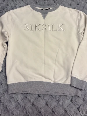 Vit sweatshirt från SikSilk - Vit sweatshirt från SikSilk med broderad logga på bröstet. Tröjan har grå ribbade muddar vid hals, ärmslut och nederkant. Klassisk rund halsringning och relaxed fit. Perfekt för en chill och stilren look. Finns lite missfärgning vid armbågarna. Se bild. 