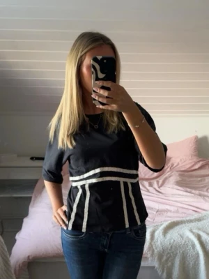 Svart topp med vita detaljer - Säljer en svart topp med vita ränder runt midjan och på sidorna. Toppen har korta ärmar och en figurnära passform. Perfekt för dig som gillar stilrena plagg med en twist.