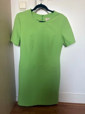 Grön kortärmad klänning - Snygg grön klänning med korta ärmar och rund halsringning. Klänningen har en enkel, rak passform och dekorativa fickdetaljer framtill. Perfekt för dig som gillar stilrena plagg med färg.