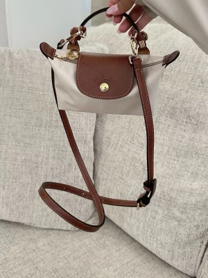 Beige och brun handväska - Beige handväska med bruna läderdetaljer och guldfärgade metalldetaljer. Väskan har ett klassiskt lock med tryckknapp och präglad logga samt ett kort handtag i läder. Axelrem tillkommer och prislappar tillkommer.💕