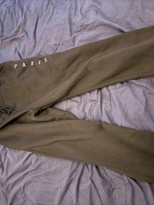 Svarta mjukisbyxor med PARIS-tryck - Säljer ett par svarta mjukisbyxor med vitt PARIS-tryck på ena benet. Byxorna har dragsko i midjan och raka ben. Perfekta för en avslappnad och trendig look. Materialet känns mjukt och bekvämt, troligen bomull eller bomullsblandning.