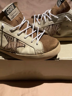 Golden Goose Superstar sneakers beige/brun - Golden Goose Superstar sneakers i beige och brunt med ikonisk stjärndetalj på sidan. Ovandel i läder och mocka, vita skosnören och högt skaft. Märkeslapp på plösen och vintageinspirerad design. Perfekta för dig som gillar streetstyle och unika detaljer.