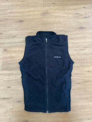 Svart fleeceväst från Patagonia - Mysig svart fleeceväst från Patagonia med hög krage och hel dragkedja framtill. Västen har en enkel och clean design med diskret logga på bröstet. Perfekt att slänga på sig över en hoodie eller tröja när det är kyligt ute.