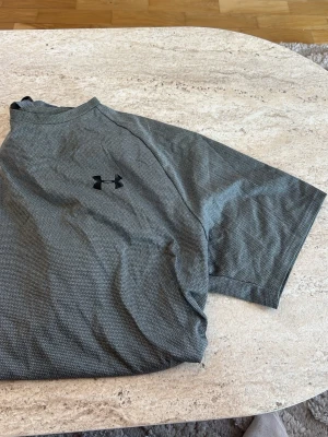 Grå träningströja från Under Armour - Säljer en grå kortärmad träningströja från Under Armour. Tröjan har diskret mönster, svart logga på bröstet och är tillverkad i ett lätt, snabbtorkande funktionsmaterial som passar perfekt till träning.