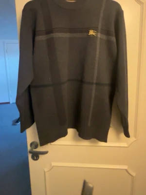 Mörkgrå stickad tröja från Burberry - Mörkgrå stickad tröja från Burberry med diskret rutmönster i olika grå nyanser och en liten guldfärgad Burberry-logga på bröstet. Tröjan har rund halsringning och långa ärmar. Perfekt för dig som gillar klassisk och stilren design.