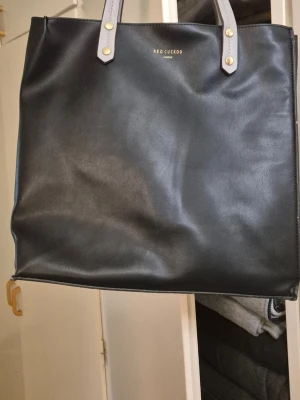 Svart shoppingväska från Red Cuckoo - Stilren svart shoppingväska från Red Cuckoo med raka linjer och rejäla handtag i ljusgrått. Väskan är tillverkad i slätt konstläder och har en enkel, klassisk design med guldfärgade detaljer vid handtagen. Perfekt för dig som gillar minimalistisk stil.