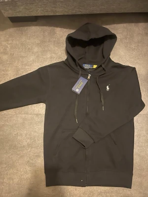 Svart hoodie från Polo Ralph Lauren - Snygg svart hoodie från Polo Ralph Lauren med dragkedja framtill och klassisk vit logga på bröstet. Hoodien har huva med dragsko och två fickor fram. Tillverkad i mjukt bomullsmaterial som är skönt att bära. Perfekt för en chill och stilren look.