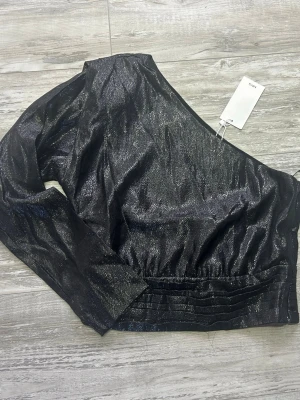 Svart metallic topp med en ärm - Snygg svart metallic topp från Mango med en axel och lång ärm. Materialet har en glansig finish och toppen har veckade detaljer vid midjan. Perfekt för dig som vill sticka ut med en unik och trendig look.