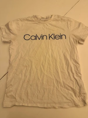 Vit Calvin Klein t-shirt i bomull - Vit t-shirt från Calvin Klein med klassisk logga tryckt i svart över bröstet. Tillverkad i mjuk bomull och har en normal passform med korta ärmar och rund hals. Perfekt basplagg för en clean och stilren look.