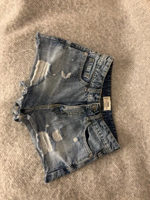 Blå slitna jeansshorts från Tally Weijl  - Säljer ett par blå jeansshorts från Tally Weijl med slitna detaljer och råa kanter. Shortsen har klassisk femficksdesign, bälteshällor och dragkedja. Perfekta för sommaren och ger en avslappnad vibe.