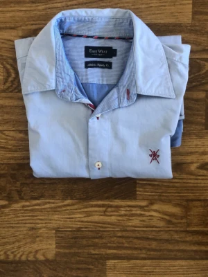 Ljusblå skjorta från East West - Klassisk ljusblå skjorta från East West med broderad detalj på bröstet. Skjortan har långa ärmar, vita knappar och en stilren krage. Perfekt för dig som gillar en clean och enkel look.