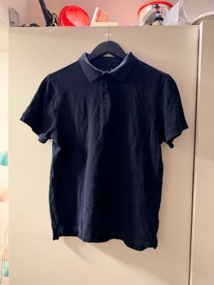 Mörkblå piké  - Klassisk mörkblå piké t-shirt storlek M med korta ärmar och krage. Tröjan har två knappar vid halsen och är tillverkad i mjukt bomullstyg. Enkel och stilren design som passar till det mesta.