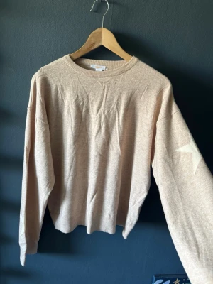 Beige stickad tröja med stjärna - Beige stickad tröja med rund halsringning och avslappnad passform. På  ärmarna finns två stora vita stjärnor som detalj, vilket är både snyggt och basic samtidigt!🤍 Tröjan har långa ärmar och ribbade muddar vid ärmslut och hals. Perfekt för dig som gillar enkla men snygga detaljer.🫶🏼🩷