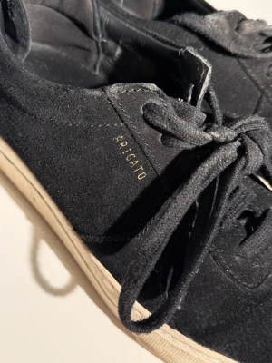 Svarta sneakers från Axel Arigato - Stilrena svarta sneakers från Axel Arigato i mocka med vit sula och klassisk låg profil. Diskret guldfärgad logga på sidan och svarta skosnören. Perfekta för dig som gillar minimalistisk och clean stil.