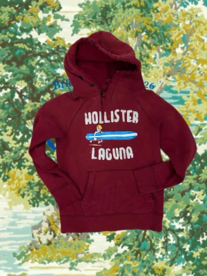 Vinröd hoodie Hollister Laguna - Snygg vinröd hoodie från Hollister med trycket 'Hollister Laguna' och en surfare på bröstet. Hoodien har huva med knappar, magficka och ribbade muddar. Perfekt för en avslappnad och cool stil. Materialet känns mjukt och bekvämt.