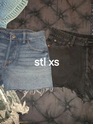 Två par jeansshorts, blå & svarta, XS - Två par jeansshorts i storlek XS. Ena paret är klassiskt blå med rå kant och femficksmodell, det andra är svart med fransig kant och slitna detaljer. Båda har hög midja och knappgylf. Perfekta för sommaren!
