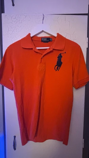 Orange pikétröja från Polo Ralph Lauren - Snygg orange pikétröja från Polo Ralph Lauren med klassisk krage och stor mörkblå logga på bröstet. Tröjan har korta ärmar och knappar vid halsen. Perfekt för en avslappnad men stilren look.